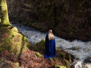 Girl with a blue cape and strawberry blonde hair, standing watching the rushing river Doon